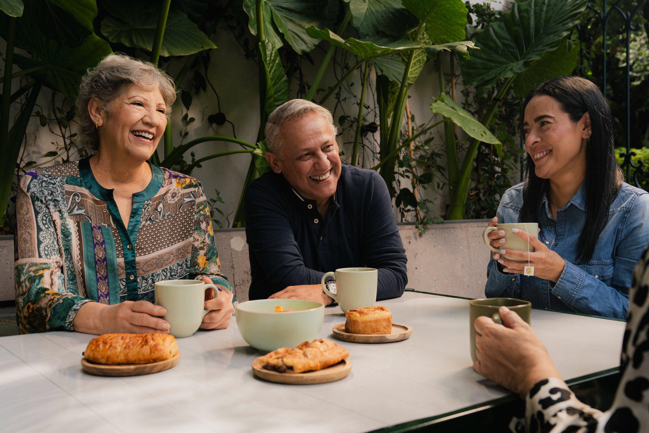 Tres personas sonriendo mientras comparten café y pan en una terraza