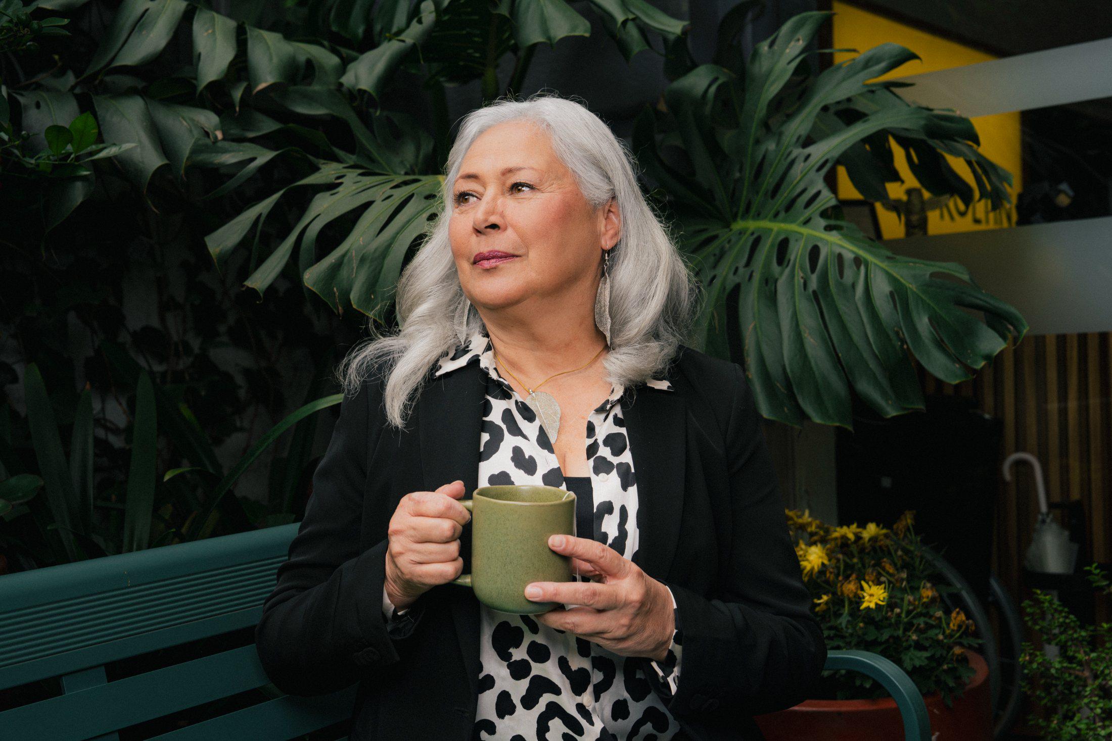Mujer de cabello cano sosteniendo una taza verde entre plantas