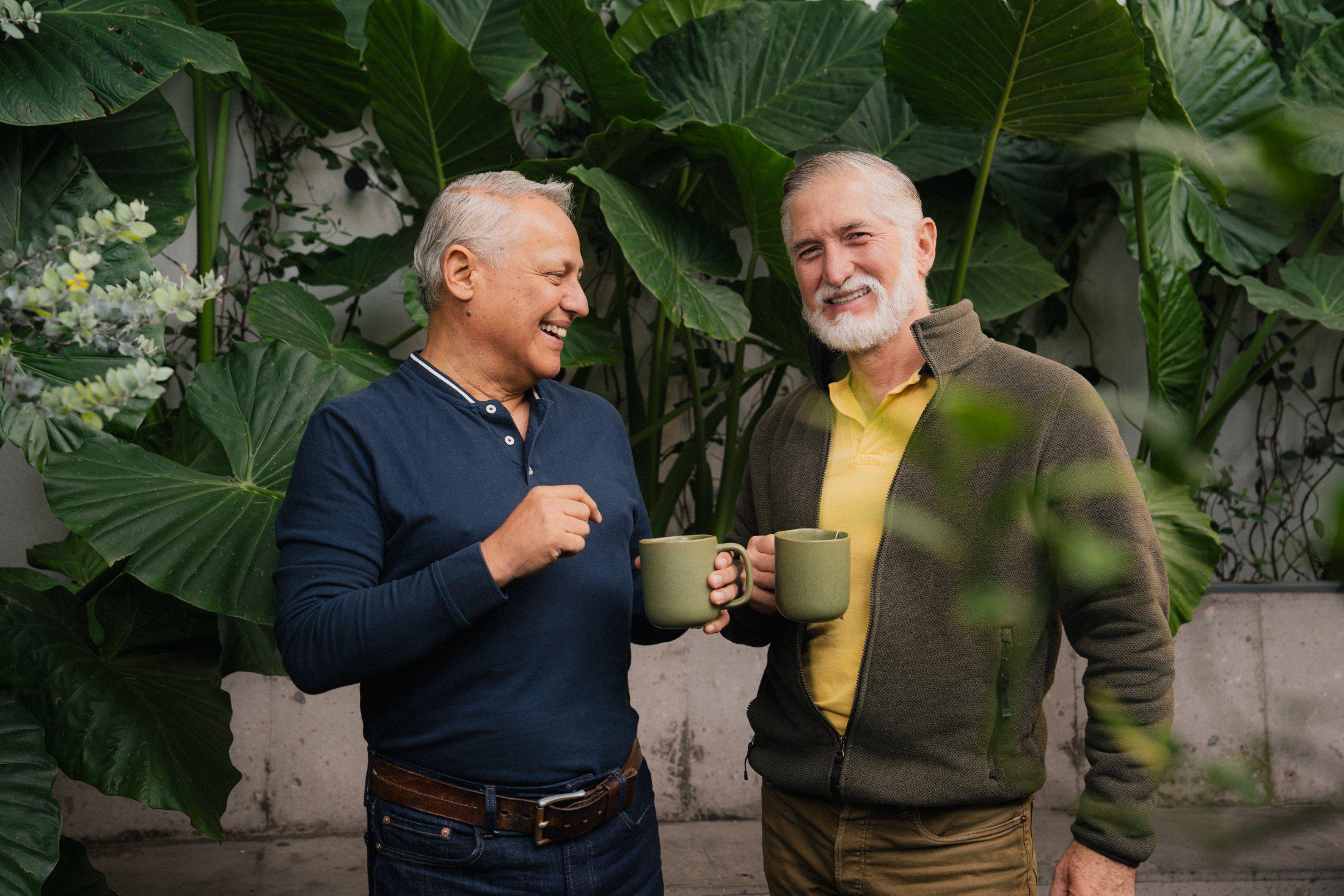 Dos hombres mayores brindando con tazas de café entre plantas